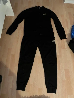 Svart EA7 loungewear set - Säljer ett svart loungewear set från EA7 med matchande ziptröja och byxor. Setet har diskreta EA7-loggor i guld på både tröjan och byxorna. Perfekt för chill dagar hemma eller när du vill vara bekvämt klädd. Mjukt material och avslappnad passform.