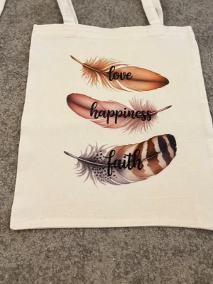 Tygkasse med fjädrar och uggla - Snygg shoppingväska i beige tyg med tryck av färgglada fjädrar och orden 'love', 'happiness', 'faith' på ena sidan och en uggla med drömfångare och blommor på andra sidan. Perfekt för dig som gillar boho-stil och unika prints. Rymlig och lätt att bära med dubbla handtag.