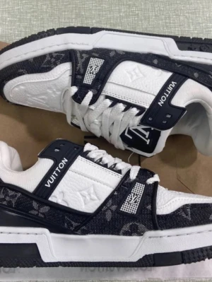 Louis Vuitton vita och svarta sneakers - Säljer ett par vita och svarta Louis Vuitton sneakers med monogramdetaljer i textil och präglat läder. Skorna har klassisk låg silhuett, vita snören och tydliga LV-loggor på sidan och tungan. Snygg kontrast mellan vitt och svart, perfekt för dig som gillar exklusiv streetwear.