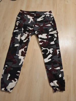 Cargobyxor i urban camo - Säljer ett par snygga  cargobyxor i Urban Camouflage. Svart, vitt, grått och vinrött. Byxorna är nya och aldrig använda! Dem har flera praktiska fickor på sidorna och baktill samt resår vid bensluten. Perfekta för en streetwear-look och är riktigt bekväma.