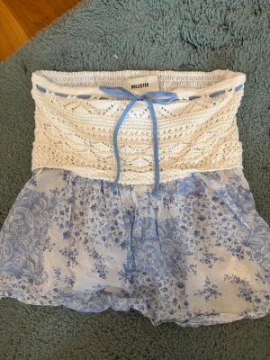 Blommig topp från Hollister - Supersöt kortkjol från Hollister med vit stickad midja och blå knytband. Kjolen har ett ljusblått blommigt mönster och volangig nederdel. Perfekt för sommarens alla tillfällen och ger en romantisk vibe.