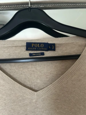 Beige stickad tröja Polo Ralph Lauren - Klassisk beige stickad tröja från Polo Ralph Lauren i mjuk pima cotton. Tröjan har v-ringning, långa ärmar och ribbade muddar. Diskret logga broderad på bröstet i mörkblått. Perfekt för en stilren och avslappnad look.