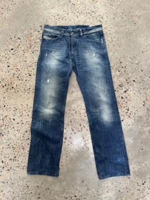 Blå raka jeans med slitningar från Diesel - Säljer ett par blå archive jeans med raka ben och snygga slitningar framtill. Jeansen har klassisk femficksdesign och en tvättad look som ger en avslappnad vibe. Perfekta för dig som gillar en casual stil och vill ha ett par jeans som funkar till det mesta. Storlek är 31/32