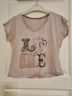 Ljusrosa t-shirt med LOVE-tryck - Söt ljusrosa t-shirt med stort dekorativt LOVE-tryck framtill i svart och vitt med blommiga detaljer. T-shirten har korta ärmar och rund halsringning. Tillverkad i mjukt bomullsmaterial och har en avslappnad passform.