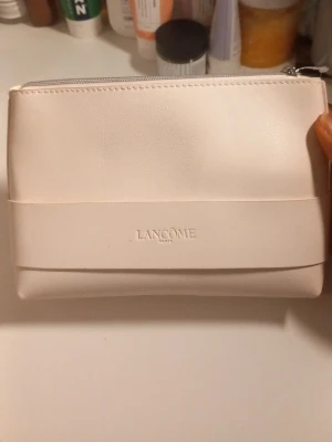 Lancôme necessär med tillagd hudvård - Säljer en stilren ljusbeige necessär i skinnimitation från Lancôme med tillagd hudvård. Necessären har dragkedja och präglad logga, krämen kommer i en lila burk med silverlock.