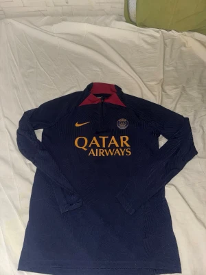 Mörkblå PSG träningströja Nike - Snygg mörkblå långärmad träningströja från PSG med gul Nike-logga och Qatar Airways-tryck på bröstet. Tröjan har halv dragkedja och vinröda detaljer vid kragen. Perfekt för fotbollsträning eller chill.
