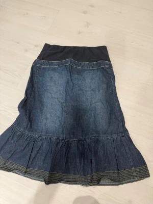Mamma  jeanskjol med knappar - Mörkblå lång jeanskjol med volangkant och dekorativa knappar framtill. Kjolen har en bred, svart midjeresår och två små fickdetaljer med knapp. Perfekt för dig som gillar en avslappnad men ändå snygg stil.