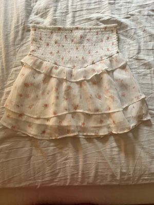 Vit blommig volangkjol - Supersöt kort kjol i vitt med rosa blommor och volangdetaljer. Kjolen har smock i midjan för en bekväm passform och dubbla lager med volanger som ger en romantisk känsla. Perfekt för vår och sommar!