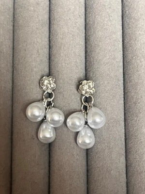 Silvriga örhängen med pärldroppar - Säljer ett par eleganta örhängen i silverfärg med gnistrande blomdetalj upptill och tre vita droppformade pärlor som hänger ned. Perfekta för dig som gillar klassiska accessoarer med en twist.