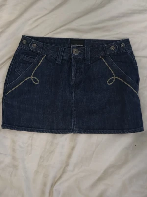 Calvin Klein kjol - Snygg mörkblå jeanskjol från Calvin Klein Jeans med klassiska fickor fram och bak, bälteshällor och kontrastsömmar. Kjolen har en kort passform och är perfekt för en trendig vardagslook.