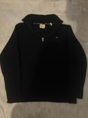 Svart halvzip tröja från GANT - Snygg mörk blå tröja från GANT med halvzip och hög krage. Tröjan har ett litet broderat GANT-emblem på bröstet och är perfekt för lager-på-lager. Klassisk och stilren design som funkar till det mesta.