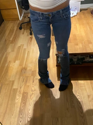 Blå slitna bootcut jeans - Säljer ett par blå bootcut jeans med slitningar och hål på knäna och låren. Jeansen har klassisk femficksmodell och låg midja.