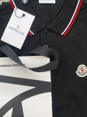 Svart pikétröja från Moncler - Snygg svart pikétröja från Moncler med klassisk krage och knappar framtill. Kragen har röda och vita ränder som ger en sportig touch. Monclers logga är broderad på bröstet. Perfekt till jeans eller shorts för en clean look.