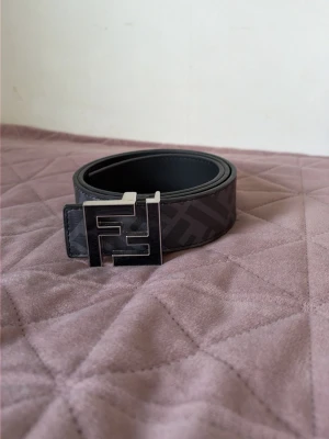 Bälte  - Snyggt svart bälte med ett stort, fyrkantigt metallspänne och diskret mönster längs remmen. Bältet har en modern design och passar perfekt till jeans eller byxor för en trendig look. Gjort i skinn för en exklusiv känsla.