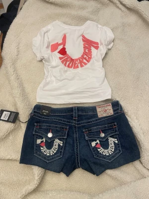Blå jeansshorts från True Religion, w29 - Strl w29, prislappen sitter fortfarande på. Säljer ett par klassiska blå jeansshorts från True Religion med broderad logga och hjärtdetaljer på bakfickorna. Shortsen har ficklock med knappar och tydliga kontrastsömmar. Perfekta för sommaren och ger en cool streetwear-vibe.