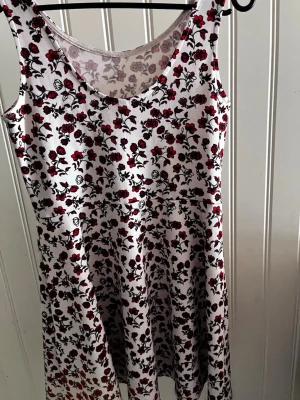 Vit blommig ärmlös kortklänning - Söt vit kortklänning med röda och svarta blommor. Klänningen är ärmlös och har en rund halsringning. Perfekt för varma dagar och har en lätt, luftig passform. Materialet känns mjukt och bekvämt mot huden.
