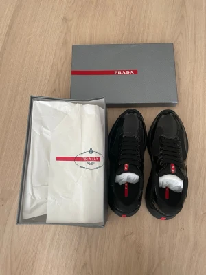 Svarta cups från Prada - Snygga svarta sneakers från Prada med glansig finish och meshdetaljer. Skorna har svarta snören och en röd logga på plösen samt hälen. Kommer i originalkartong med silkespapper. Perfekta för dig som vill ha en clean och exklusiv look.