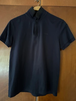 Hugo Boss piké - Mörkblå med svarta kontrast detaljer. Använd fåtal gånger, säljes pga för liten. Mycket bra skick. Storlek S. 92% bomull 8% elastan.
