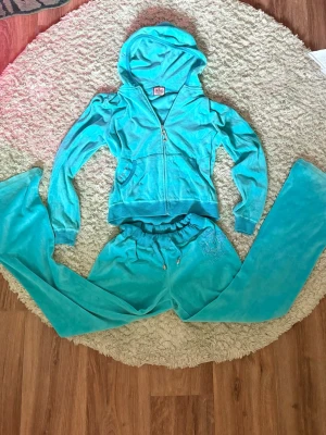 Turkost Juicy Couture velourset från 2000s - Säljer ett ikoniskt och ovanligt turkost velourset från Juicy Couture. Setet består av en zip-hoodie med fickor och huva samt matchande byxor med resår i midjan. Baksidan av hoodien har Juicy Couture-logga och glittriga detaljer. Perfekt för chill dagar hemma. Från 2000s. 