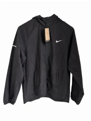 Svart vindjacka från Nike med huva - Snygg svart vindjacka från Nike med vit logga på bröstet och dragkedja framtill. Jackan har huva, två sidofickor och elastiska muddar vid ärmsluten. Tillverkad i lätt och vindtätt material, perfekt för blåsiga dagar.