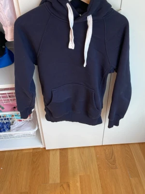 Russemerch - En stilren mörkblå hoodie med klassisk känguruficka och vita dragsnören i huvan. Hoodien har ribbade muddar vid ärmar och nederkant samt en mjuk insida.