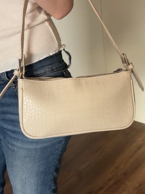 Beige axelremsväska i crocopräglat skinn - Snygg beige handväska med axelrem och crocopräglat mönster. Väskan har en avlång, rektangulär form och stängs med dragkedja. Perfekt för dig som gillar stilrena accessoarer med trendig känsla.