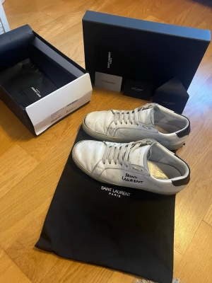 Vita Saint Laurent skor - Snygga vita låga sneakers från Saint Laurent i äkta läder med klassisk siluett. Skorna har svarta detaljer på hälen, vita skosnören och Saint Laurent-logga på sidan och plösen. Perfekta för dig som gillar stilrena och exklusiva sneakers.