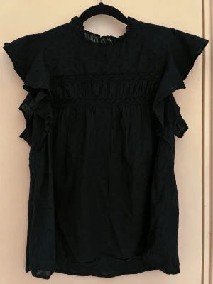 Svart blus med volang - Svart blus med volangdetaljer vid ärmarna. Blusen har ett prickmönster i tyget och dekorativa sömmar över bröstet. 