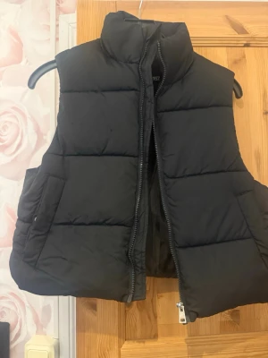 Svart puffad dunväst med hög krage - Snygg svart dunväst med puffad design och hög krage. Västen har dragkedja framtill och två praktiska sidofickor. Perfekt för lager-på-lager och ger en cool streetwear-look. Passar till både hoodie och t-shirt.