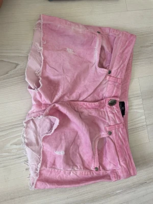 Ljusrosa jeansshorts med fransig kant - Säljer ett par ljusrosa jeansshorts med fransig rå kant och slitna detaljer. Shortsen har klassisk femficksdesign, knappgylf och en avslappnad passform. Perfekta för sommardagar och festivaler.