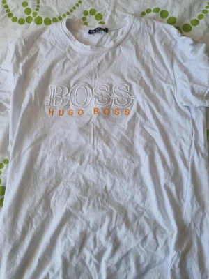 Vit kortärmad t-shirt från Hugo Boss - Säljer en vit t-shirt från Hugo Boss med broderad logga i vitt och orange text på bröstet. Klassisk rund halsringning och korta ärmar. Tillverkad i mjuk bomull för en skön känsla och enkel stil.