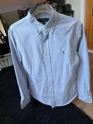 Blårandig skjorta från Polo Ralph Lauren - Klassisk blårandig skjorta från Polo Ralph Lauren med button-down krage och broderad logga på bröstet. Skjortan har långa ärmar, knappar framtill och är gjord i mjuk bomull. Perfekt för en clean och stilren look.