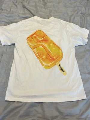 Vit Stussy t-shirt med glassmotiv - Vit t-shirt från Stussy med ett stort, färgglatt glassmotiv på ryggen och liten Stussy-logga framtill. T-shirten har klassisk passform och korta ärmar. Perfekt för dig som gillar streetwear och unika prints. pris går att diskutera