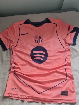 Fotbolls T-Shirt - Säljer en rosa FC Barcelona t-shirt med mörkblå detaljer och tryck. Tröjan har korta ärmar, rund hals och tryck med Spotify-logga fram samt Raphinha och nummer 11 bak. Tillverkad i ett lätt och funktionellt material, perfekt för fotboll eller träning.