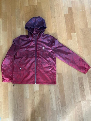 Vinröd vindjacka med huva - Snygg vinröd vindjacka från Thrasher med huva och dragkedja framtill. Jackan har två fickor med knappar, justerbar nederkant och elastiska ärmslut. Thrasher-logga på bröstet och stor text på ryggen. Materialet är lätt och vattenavvisande.