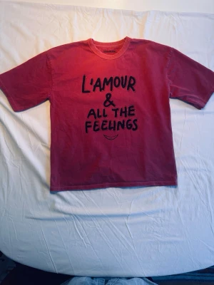 Röd t-shirt med tryck L'amour - röd tvättad t-shirt med svart texttryck 'L'amour & all the feelings' på framsidan. Klassisk rund halsringning och korta ärmar. Perfekt för dig som vill ha en statement-tee med skön passform. Märke Zadig & Voltaire storlek 14-S