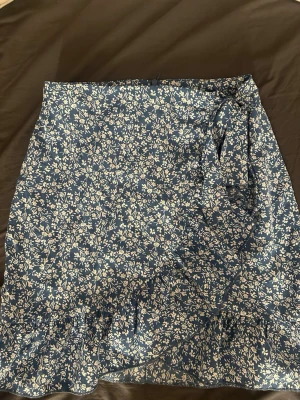 Blå blommig omlottkjol med volang - Söt blå omlottkjol med vitt blommönster och volangkant. Kjolen har knyt i sidan och är i ett lätt, mjukt material som passar perfekt till varmare dagar. Den har en kort längd och ger en avslappnad men ändå trendig look.
