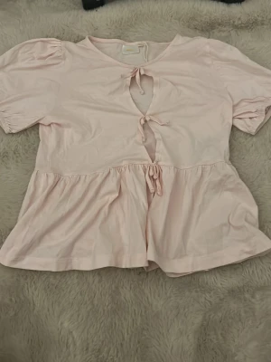 Ljusrosa topp med knyt och volang - Söt ljusrosa topp med korta puffärmar och två knyt framtill som skapar öppningar. Toppen har en volangkant nedtill som ger en avslappnad och feminin look. Tillverkad i ett mjukt, tunt tyg som känns skönt mot huden.
