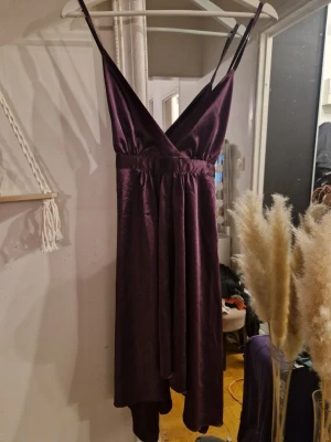 Mörklila långklänning med smala band - Säljer en elegant mörklila långklänning i glansigt material med smala axelband och v-ringning. Klänningen har omlott-look framtill och justerbara band i ryggen. Perfekt för dig som vill ha en stilren och chic look. Dragkedja i sidan. 