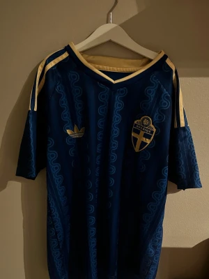 Blå Sverige fotbollströja Adidas - Säljer en blå fotbollströja från Adidas med Sveriges landslagsemblem på bröstet och gula detaljer på axlar och krage. Tröjan har ett mönster i tyget och texten 'Sverige' på ryggen. Perfekt för match eller träning.