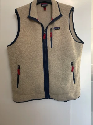 Beige fleeceväst från Patagonia - Säljer en beige eftertraktad fleeceväst från Patagonia med marinblå detaljer och röda dragkedjor. Västen har tre fickor med dragkedja och en hög krage. Perfekt att slänga på sig över en hoodie eller tröja för extra värme särkilt nu till hösten/sen sommar🙌