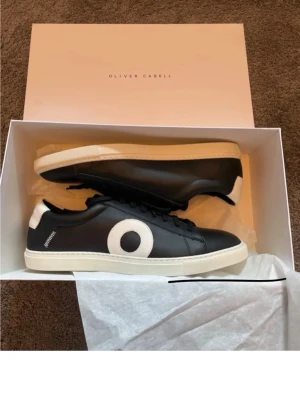 Svarta sneakers från Oliver Cabell - Stilrena svarta sneakers från Oliver Cabell med vit sula och vit detalj i form av en cirkel på sidan. Skorna har svarta skosnören och vit hälflik. Tillverkade i slätt läder för en clean look. Perfekt nu till sommaren. Hör av er vid funderingar!