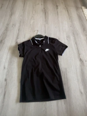 Svart pikétopp från Nike - Säljer en svart pikétopp från Nike med vit logga på bröstet och vita detaljer på krage och ärmslut. Klassisk polokrage med knapp och korta ärmar. Perfekt för en sportig och stilren look.