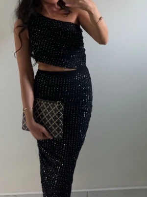 Svart strass oneshoulder set - Snyggt svart oneshoulder set med strass. Klänningen har en åtsittande passform och är croppad upptill, vilket ger en trendig look. Perfekt för dig som vill sticka ut med stilrena detaljer och en festlig känsla. Passar även xs. 🪩