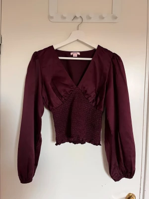 Vinröd blus med v-ringning - Snygg vinröd blus med djup v-ringning och smockad midja. Blusen har långa puffärmar och är tillverkad i ett glansigt, mjukt tyg som ger en elegant känsla. Perfekt att styla med både jeans och kjol för en trendig look.