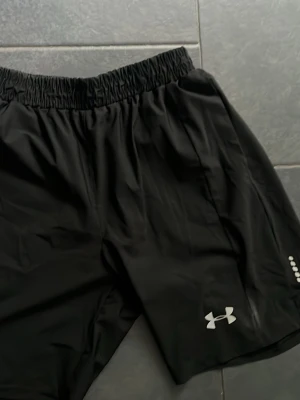 Svarta träningsshorts från Under Armour - Snygga svarta träningsshorts från Under Armour med elastisk midja och logga i vitt på ena benet. Tillverkade i lätt och snabbtorkande syntetmaterial, perfekta för gymmet,  löprundan eller om du bara vill gå runt i snygga shorts. Shortsen passar även L