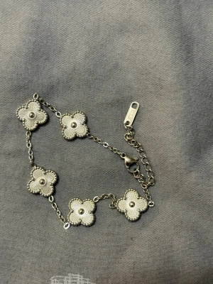 Silverfärgat armband med blommor - Ett stilrent armband i silverfärg med fem dekorativa blommor i rad. Blommorna har detaljerade kanter och en liten pärla i mitten. Armbandet har justerbar kedja och karbinlås. Perfekt för dig som gillar unika och eleganta accessoarer. Vattenfast 