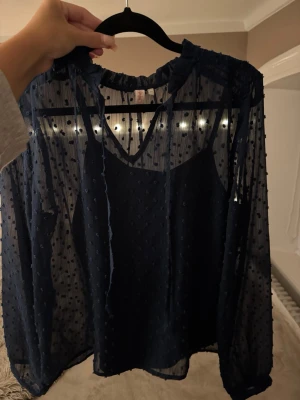 Mörkblå transparent blus med prickar - Snygg mörkblå blus i transparent material med små prickar och volangdetaljer vid halsen. Blusen har långa ärmar och medföljande linne under. Perfekt för dig som gillar trendiga och unika plagg.