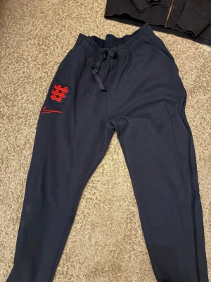 Mörkblå Nike England Dri-FIT byxor - Säljer ett par mörkblå träningsbyxor från Nike med England-logga och röd Nike swoosh på vänster ben. Byxorna har dragsko i midjan och är tillverkade i Dri-FIT-material som andas. Perfekta för träning eller chill.