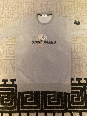 Grå hoodie från Stone Island - Snygg grå hoodie från Stone Island med klassisk logga broderad på bröstet och ikonisk patch på ärmen. Hoodien har rund halsringning och ribbade muddar vid ärmslut och nederkant. Perfekt för en stilren och avslappnad look.